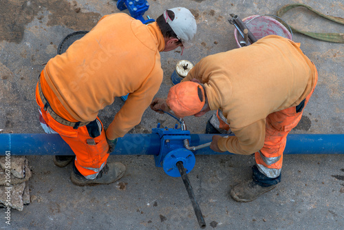 Overhead view of two plumbers repairing a blue water pipeline on a construction site. Workers in orange safety uniforms fixing a pipe connection with wrenches. Commercial industrial stock.