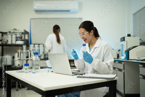 Excited Asian Female Scientist Examining Test Tube in Modern Laboratory with Laptop and Lab Equipment