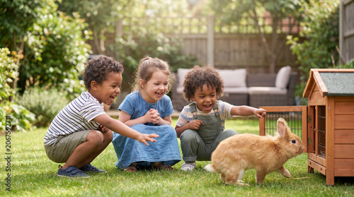 Three children playing with a brown rabbit in a green garden, wooden rabbit hutch visible, surrounded by plants and a cozy seating area in the background