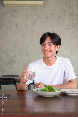 Young Asian non-binary man smiling holding glass of water looking away portrait