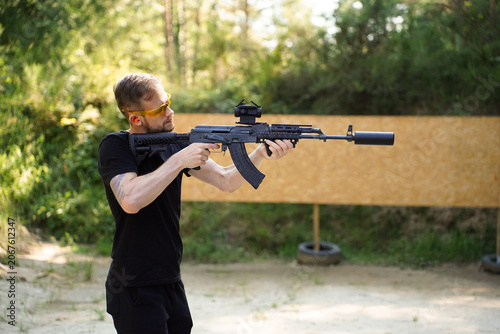 Slika na platnu Man in black shirt practicing tactical shooting with rifle and suppressor at outdoor firing range