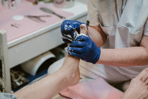Feet care, a pedicurist wearing blue gloves for hygiene is performing foot care on a female client inside a feet spa clinic.