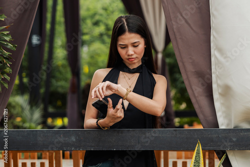 Foto A young Southeast Asian woman checks her wristwatch while holding a smartphone and waiting in an outdoor alfresco area