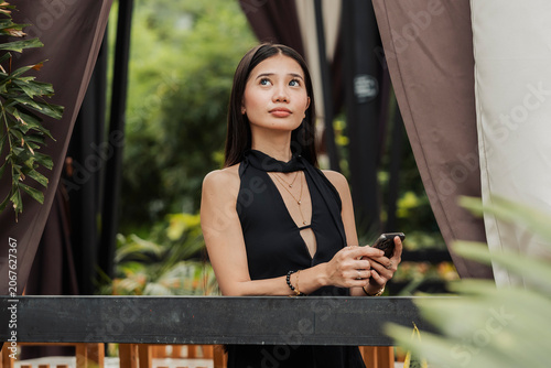 Canvastavla A young Southeast Asian woman looks up after checking a weather report on her smartphone in an outdoor alfresco setting