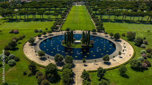 Photos Aerial view of a large circular memorial pond with fountains and an island of cypress trees, leading into a long green lawn surrounded by woods
