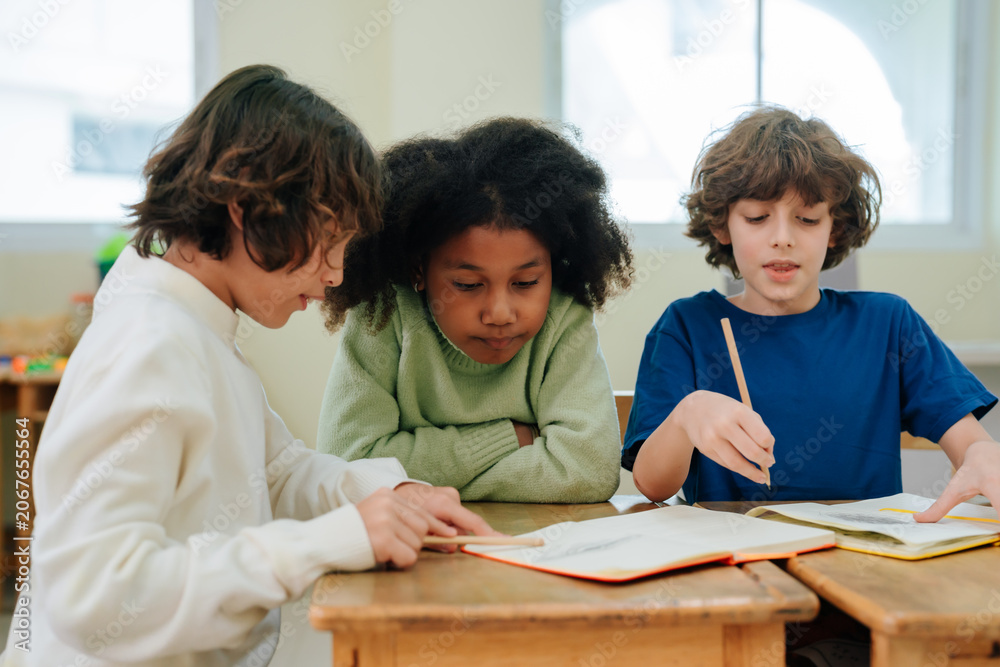 Fototapeta premium Diverse students sitting at classroom desk reading book and writing with pencils during school lesson within educational environment