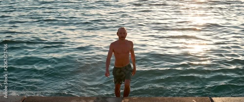 Man walking out of the sea at sunset, climbing stone steps with wet skin and warm backlight, anamorphic static shot.
