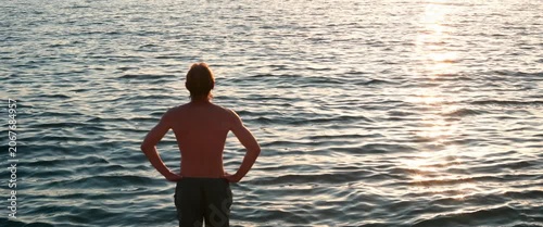 Man standing calmly in the sea at sunset, gazing at the horizon with golden sunlight reflecting on water, anamorphic static shot.
