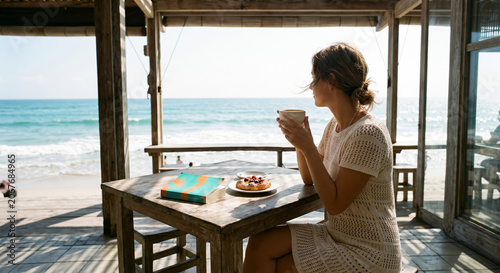 Side profile of a woman enjoying a warm drink at a rustic wooden table overlooking a serene ocean view.