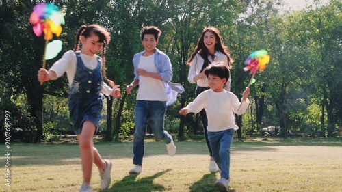 Happy Asian family with parents, daughter, and son holding colorful pinwheels while running and playing together on the grass in a sunny park, representing a healthy lifestyle and wellness