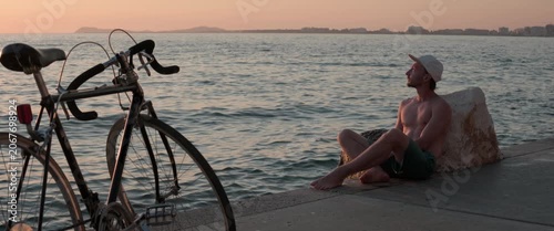 Man sitting on a pier watching the sunset, with a bicycle parked nearby and a coastal city skyline, anamorphic static shot.