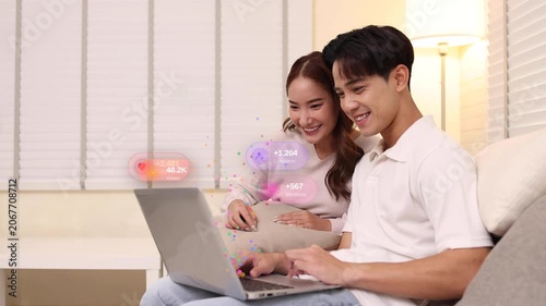 Happy young Asian couple content creator sitting on a couch, looking at laptop with floating social media notification icons like likes, comments, shares, showing viral success and digital marketing