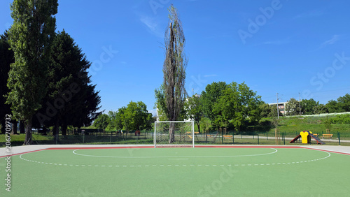 White metal soccer goal with net on a green hardcourt sports field bordered by trees under a blue sky