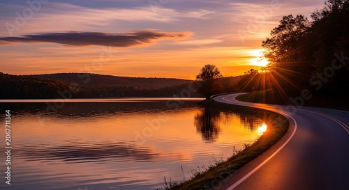 Winding road hugs tranquil lake under vibrant sunset sky, trees reflect