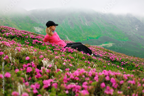 Canvas-taulu closeup of hiking girl siting on hill with beautiful wild rhododendron pink flowers on mountain peak with clouds in the background during summer time