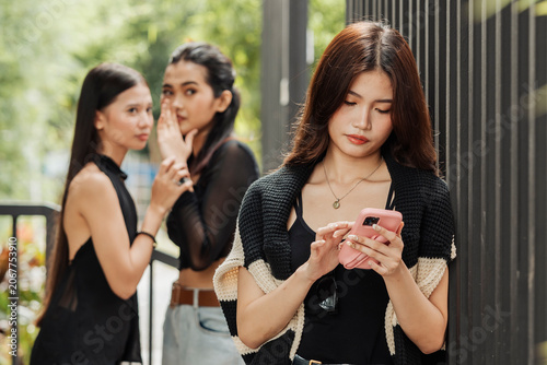Photography Two girls whisper and gossip behind a woman focused on her phone, capturing social judgment, exclusion, and subtle tension