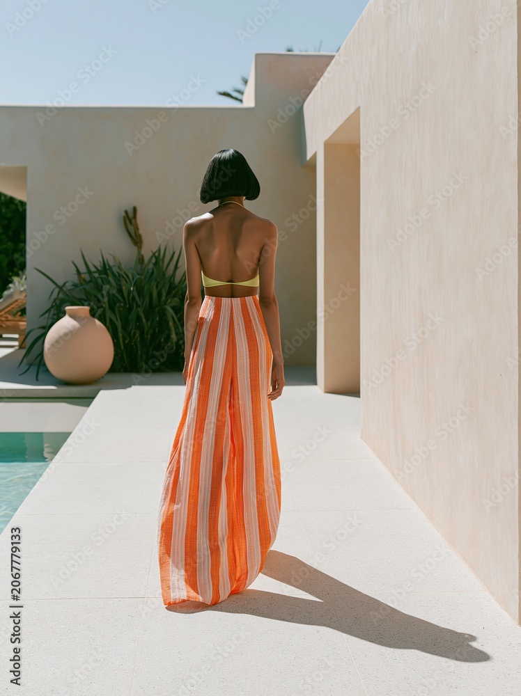 Obraz premium Woman in a striped maxi dress walks away from the camera beside a swimming pool, with a modern architectural backdrop and greenery in the scene