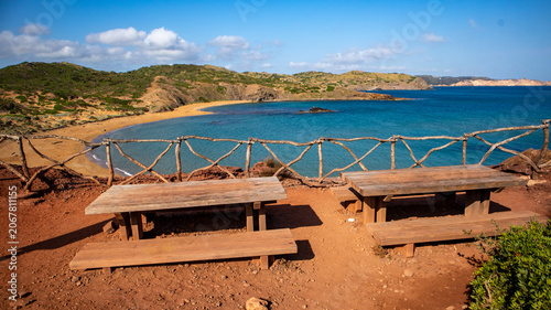 Two wooden picnic tables overlooking a scenic bay with a sandy beach and hills