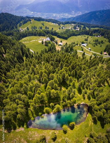 Aerial drone view of the beautiful Lago delle Prese in Valsugana, featuring an emerald alpine lake, lush coniferous forest, and rustic mountain cabins in Trentino, Italy