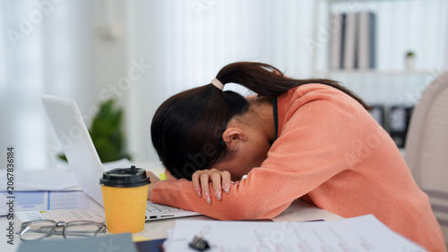 Exhausted Asian businesswoman resting head on desk beside laptop and coffee expressing work stress at home office