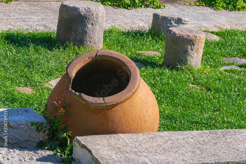 Ancient clay storage vessel displayed outdoors among stone artifacts and green grass in archaeological setting