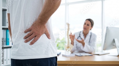 Man suffering from back pain consulting with female doctor in office while she explains spinal condition using anatomical skeleton model