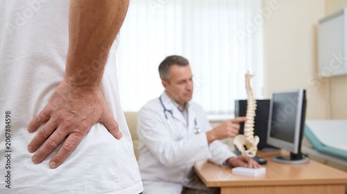 Patient having consultation with male doctor about back pain using anatomical spine model in modern medical clinic office setting