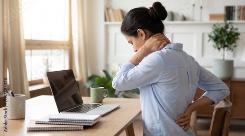 Stressed young woman working at home office suffering from neck and lower back pain while sitting at desk