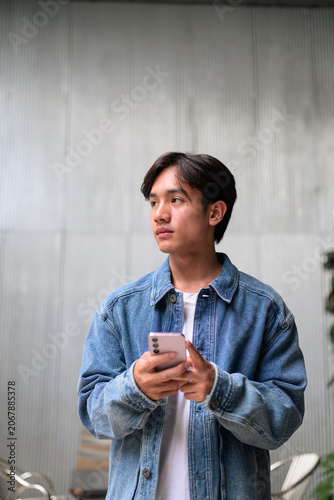 Young Asian non-binary man holding phone looking away by corrugated wall