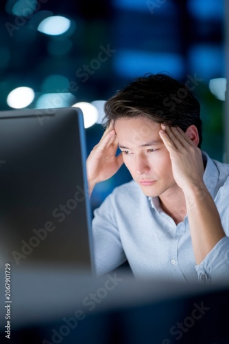a tired business person working late at night in the office, hands on their head and looking into a computer monitor