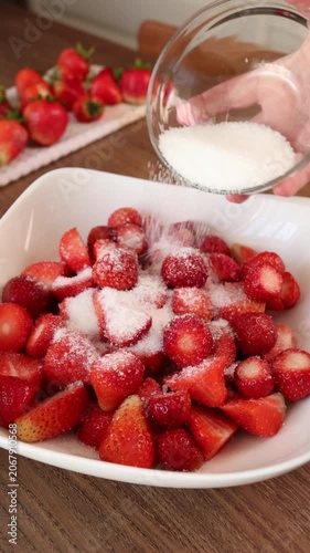 Pouring white sugar from glass bowl onto fresh cut strawberries in white plate. Sweet homemade food preparation on wooden kitchen table reflecting traditional rustic recipe. Authentic trends in simple