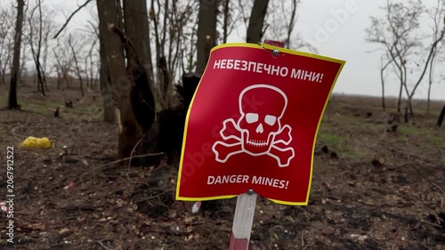 Red skull-and-crossbones mine-warning sign on a marker stake at the edge of a war-damaged forest of charred, shrapnel-scarred trees under an overcast late-autumn sky