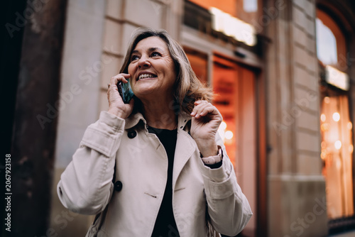 Smiling mature woman talking on smartphone in evening city, middle aged traveler using cellular roaming near historic building, authentic representation of digital nomad lifestyle and connection.
