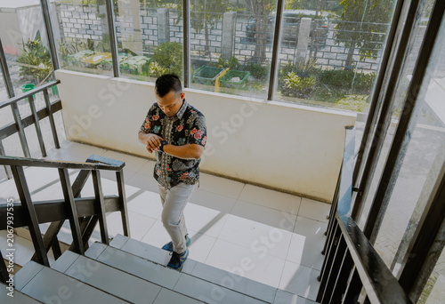 Confident adult Asian businessman in batik clothes culture looks her wristwatch and walking on building office stairs interior.