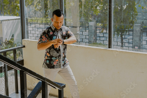 Confident adult Asian businessman in batik clothes culture looks her wristwatch and walking on building office stairs interior.