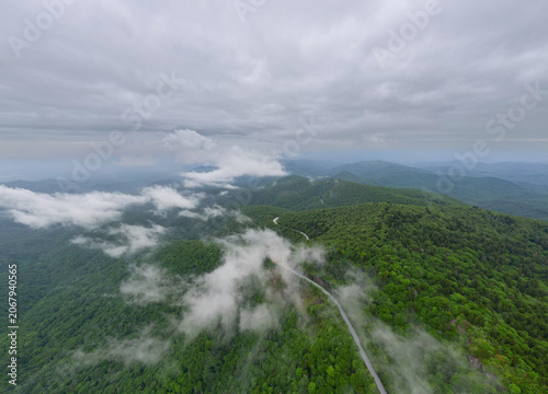Drone perspective of the Blue Ridge Parkway stretching into the distance, flanked by dense green forests, with scattered mist drifting above the treetops in the Appalachian mountains