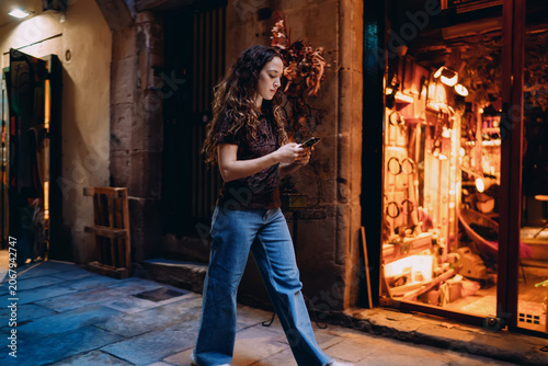 Fotografia Focused young woman walking while browsing phone, immersed in digital task, blending personal mobility and modern tech use on city street