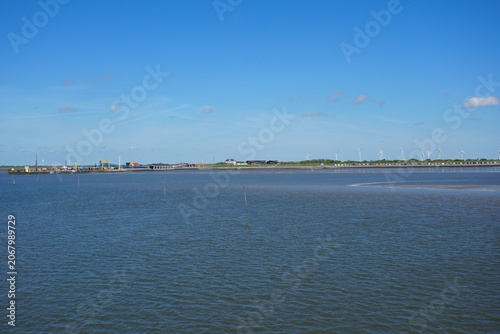 A scenic view from a ship approaching the coastline of Dagebüll, North Friesland, Germany, showing the harbor, colorful beach huts and wind turbines on the horizon under a clear sky