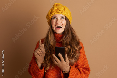 Cheerful woman in yellow beanie and orange sweater smiles broadly, looking upwards with index finger pointed, holding smartphone. She embodies eureka moment of inspiration or exciting new idea