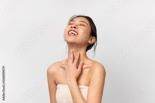 Fotografie Happy Asian Woman Smiling While Applying Skin Care Product On Her Neck At Home, Enjoying Self-care Routine