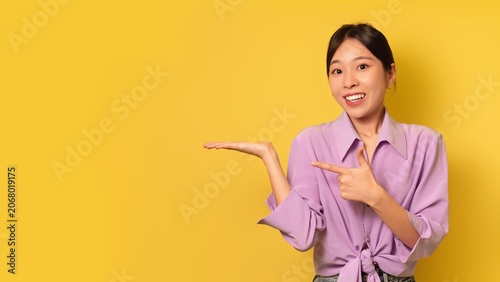 фотографія Smiling Asian Woman Pointing At Empty Space With Happiness, Showing Excitement And Enthusiasm, Standing In Front Of Bright Yellow Background, Suitable For Advertising And Presentation Concept