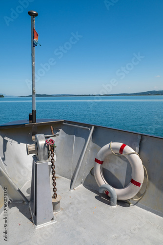 Sunny view from a small boat bow with lifebuoy, calm blue lake water, and distant shoreline under clear sky.