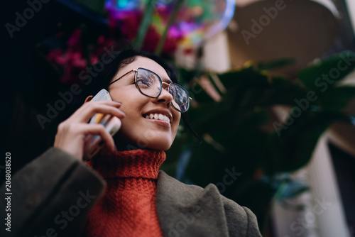 Tableau sur toile Close up of happy mixed race woman talking on cellphone, wearing glasses and orange turtleneck, urban setting, joyful emotion, wireless communication, mobile app user, authentic lifestyle