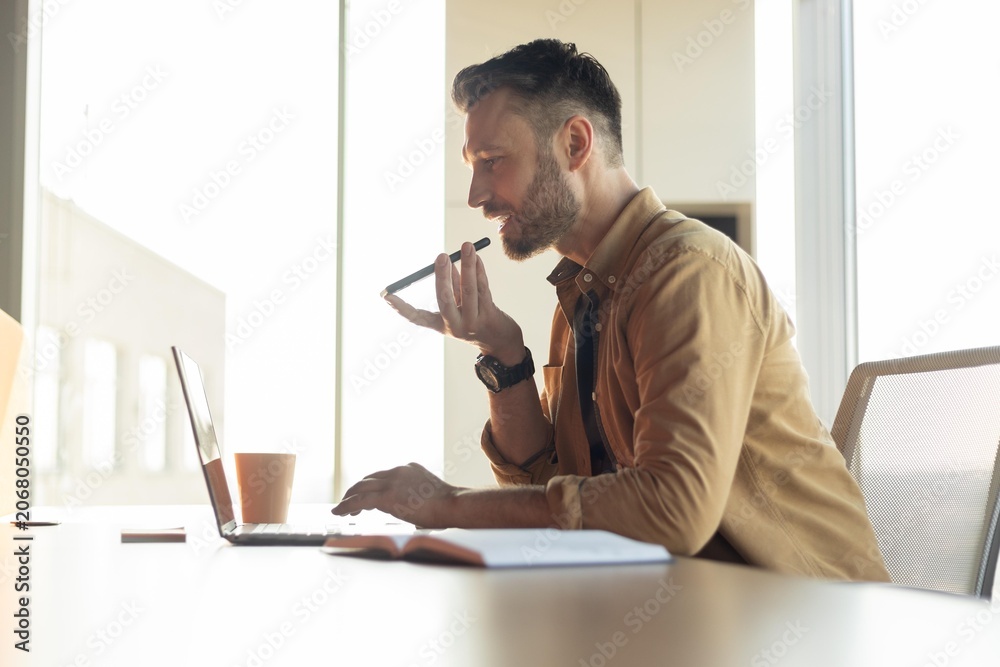 Fototapeta premium Focused Caucasian Male Working From Home, Using Laptop And Voice Recorder While Sitting At Desk In Modern Office. Professional Atmosphere With A Concept Of Remote Working And Productivity.