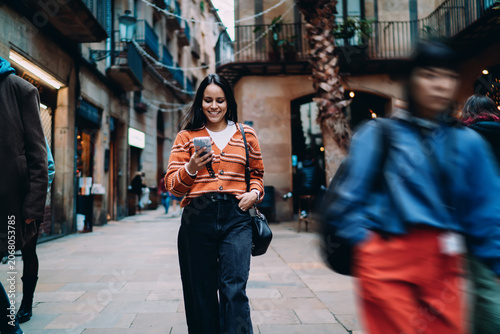 Smiling Hispanic woman walking on city street using smartphone. Young dark skinned model in orange cardigan browsing social media. Authentic representation of digital lifestyle and urban fashion.
