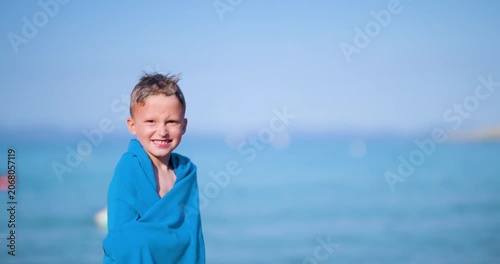 Happy little boy with wet hair and a big smile, wrapped in a bright blue towel, joyfully jumps by the clear blue ocean under a bright summer sky, enjoying his beach vacation.