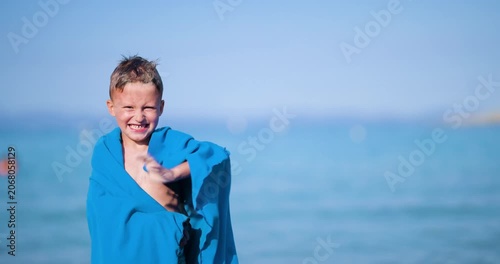Happy little boy wrapped in a blue towel smiles and waves at the camera while enjoying a sunny day at the beach with the beautiful blue sea in the background.