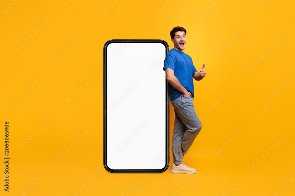 Fototapeta premium Happy Male Appearing Excited While Standing Beside A Large Smartphone With An Empty Screen, In A Bright Studio Setting. Promoting New App Or Digital Service, Advertising Concept.