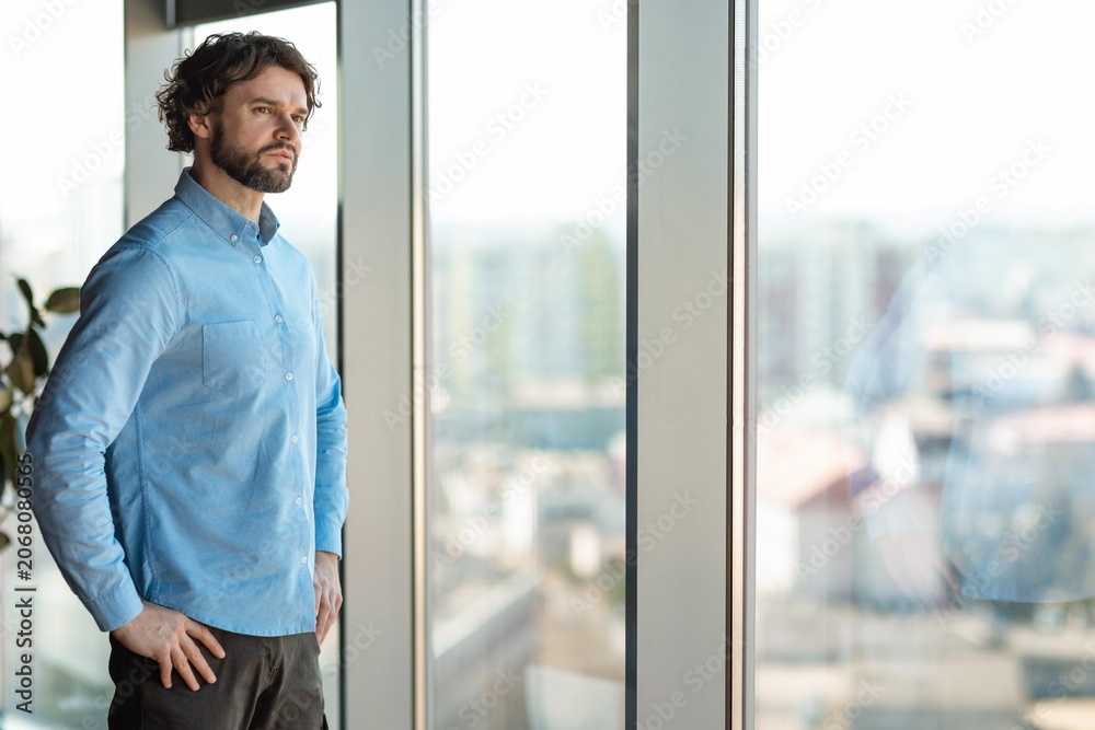 Fototapeta premium Focused Man In Mid-30s, Caucasian, Contemplating While Looking Out Large Window In Contemporary Office Space, Reflecting On Challenges And Opportunities. Business Concept.