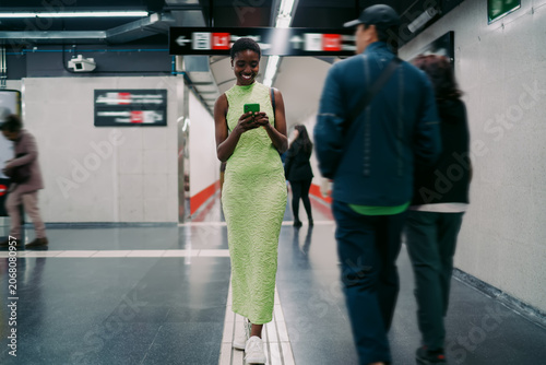 Foto Smiling black woman in neon dress text messaging on smartphone inside underground subway station